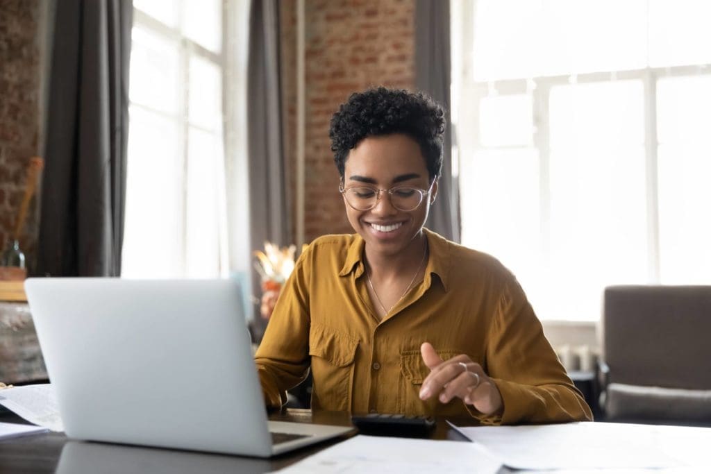 Woman sitting in front of a laptop with a calculator, smiling in a sunny room. 