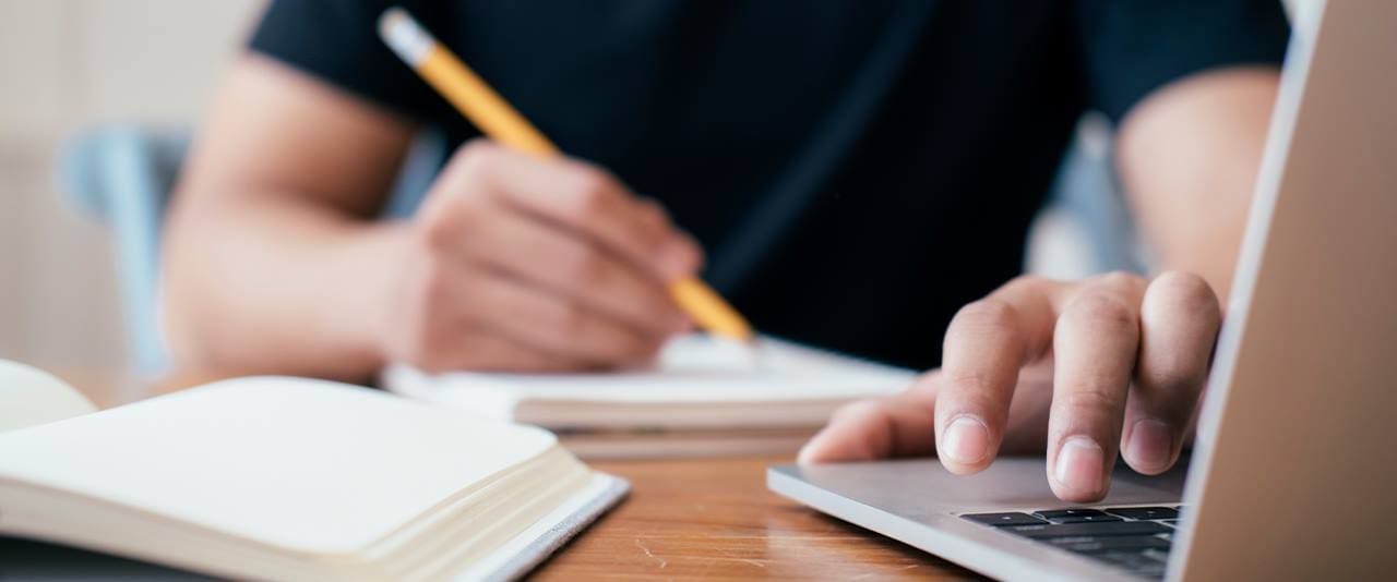 Hands using a pencil to write in a notebook and a laptop. 