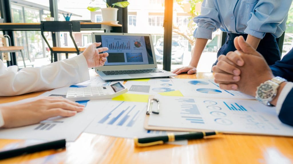 Papers and pens all scattered over a large desk while business men and woman discuss an initiative