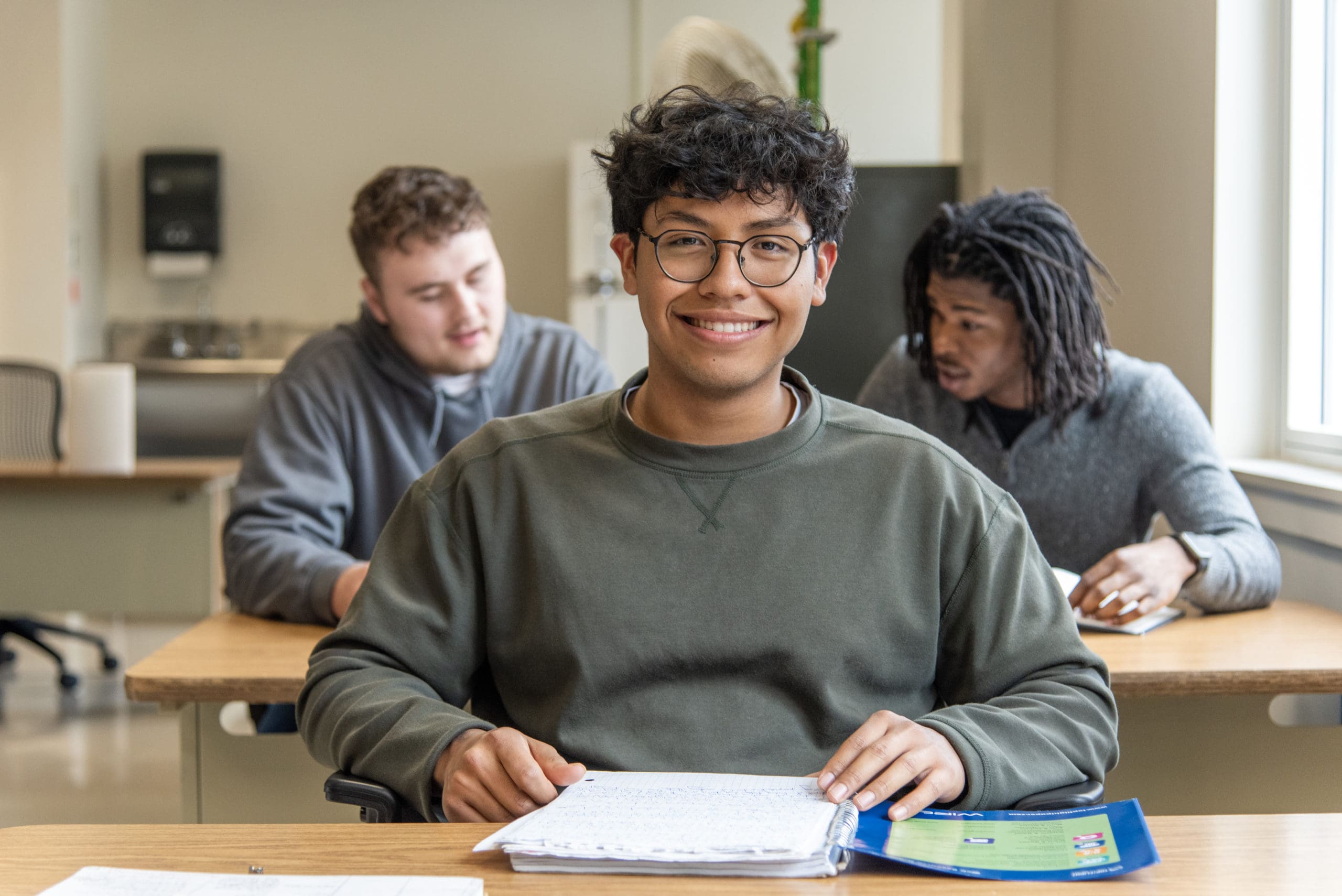 Thaddeus Stevens College of Technology student wearing glasses smiling at desk with two peers in the background working together and laughing